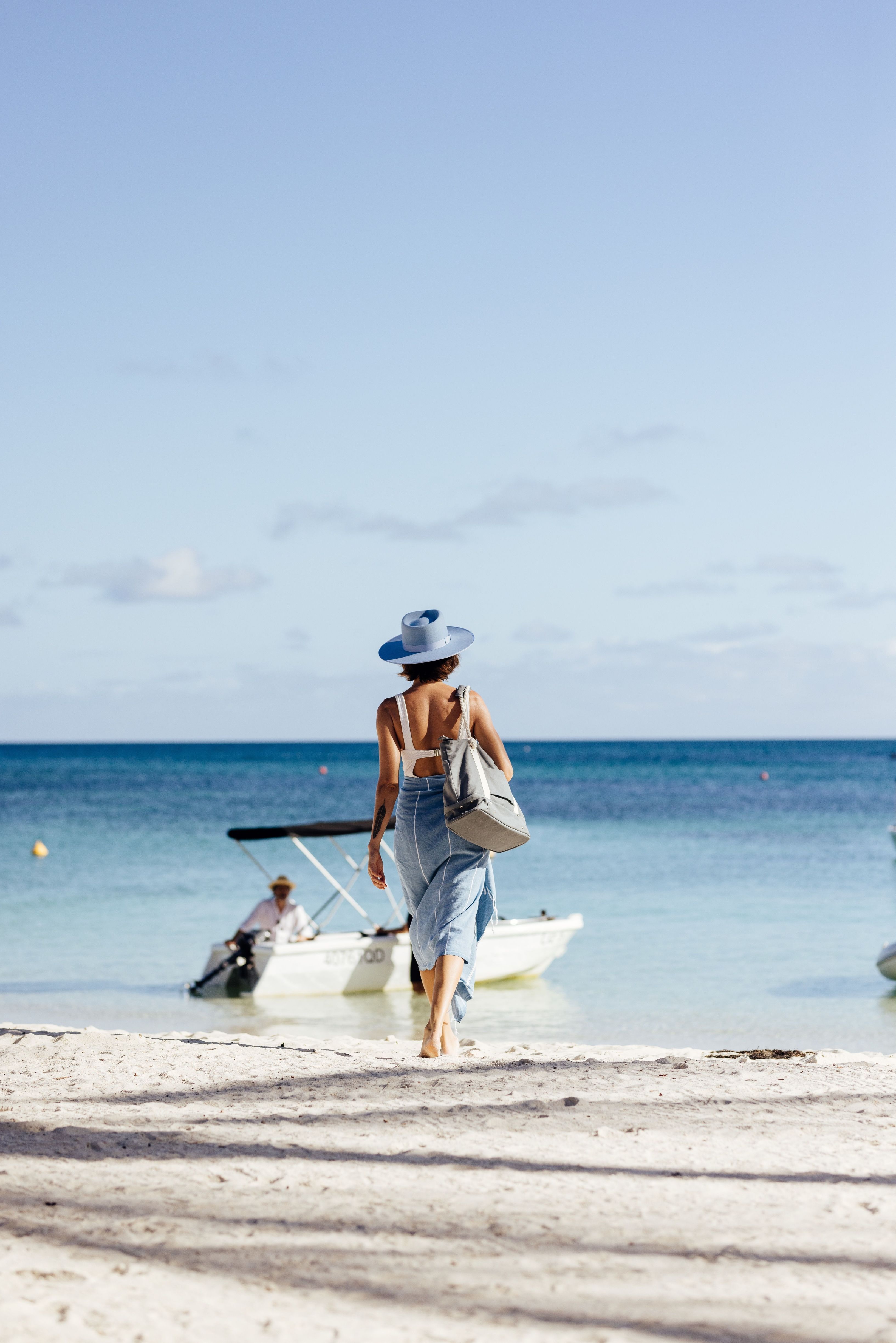 A woman wearing a hat and sarong walks across white sand towards turquoise water and a small boat at Lizard Island, Great Barrier Reef in sunny Tropical North Queensland. © Tourism Australia/Luxury Lodges of Australia
