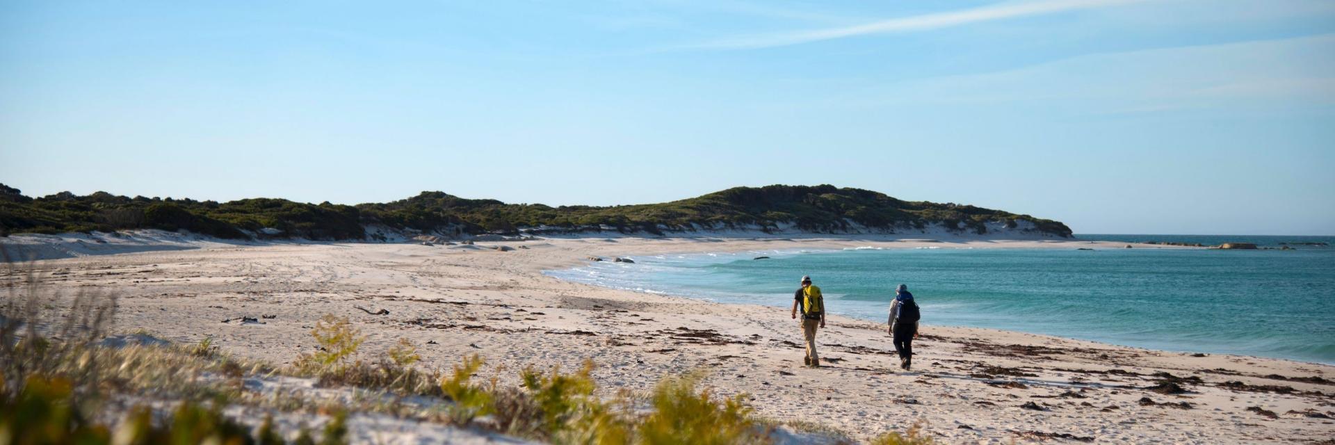 Two hikers walk along the pristine white sands of a secluded beach on the wukalina Walk in the Bay of Fires, Tasmania. Turquoise waters lap the shore, bordered by low coastal scrub and a clear blue sky on the horizon. © The wukalina Walk