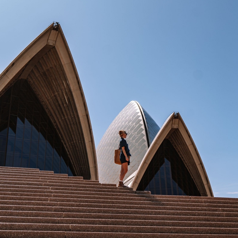 A person stands at the pinnacle of the steps that ascend to the iconic Sydney Opera House. Captured from a low angle, the image emphasises the contrasting geometric patterns: the stark, straight lines of the steps juxtaposed with the sweeping, curved sails of the Opera House. The composition is both dramatic and artistic, highlighting the grandeur of the architecture and the solitary figure's moment of contemplation against the monumental backdrop. The clear sky frames the scene, adding to the overall sense of openness and scale. © Tourism Australia