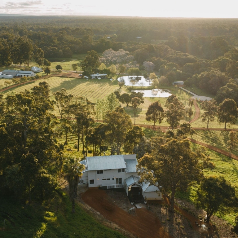 An aerial view of the picturesque Ampersand Estates in Pemberton, Western Australia, captured at sunset. The white farmhouse is surrounded by lush green vineyards, rolling hills, and tranquil dams reflecting the golden afternoon light. © Tourism Australia