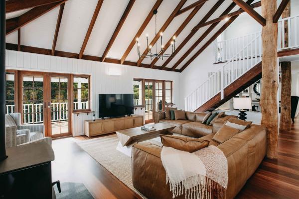 The stylish interior of luxury accommodation at Ampersand Estates in Pemberton, Western Australia. The living area features high vaulted ceilings with exposed timber beams, a large leather sectional sofa, and a rustic log pillar that adds a touch of wilderness charm to the modern décor. © Tourism Australia