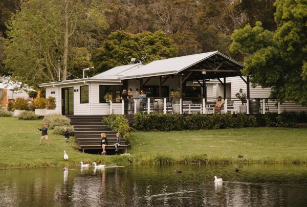 Children play on the lawn and steps by a tranquil pond at Ampersand Estates, Pemberton, Western Australia. Guests relax on the large covered deck of the white weatherboard cellar door, while ducks and geese glide across the water in the foreground. © Tourism Australia