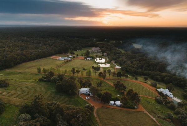 A wide aerial shot captures Ampersand Estates in Pemberton, Western Australia, during a dramatic sunset. The sprawling estate features vineyards, dams, and white farmhouses bordered by a vast, dense forest. © Tourism Australia