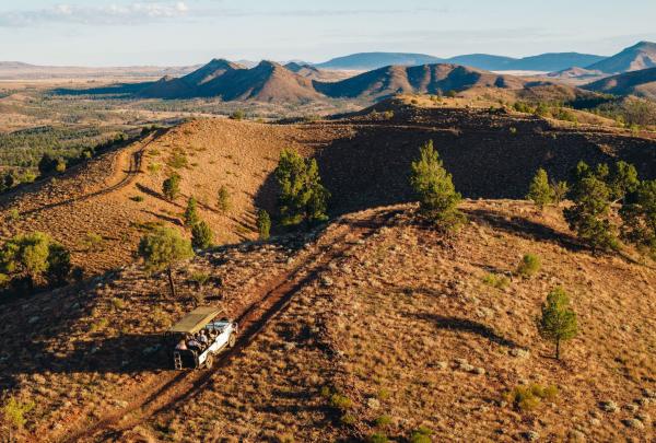 An open-top safari vehicle navigates a winding dirt track along a rugged, sun-drenched ridge at Arkaba, Flinders Ranges. Sparse green trees dot the ancient, rocky orange landscape that stretches toward layered blue mountains under a soft morning sky. © The Arkaba Walk & Great Walks of Australia