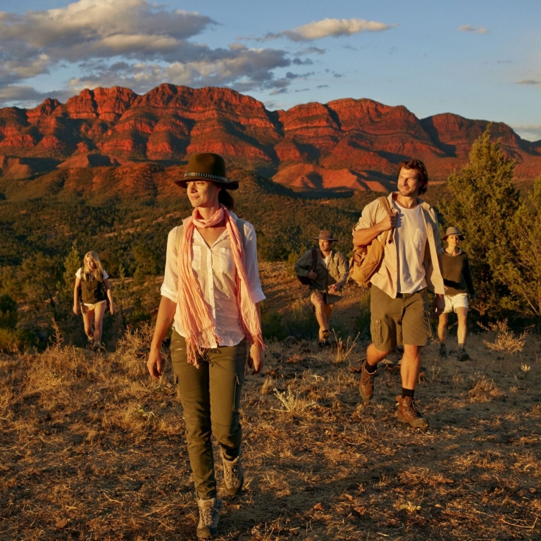 A group of guests enjoys a guided hike through the rugged terrain of Arkaba, Flinders Ranges, South Australia. Clad in outdoor gear, they walk across a grassy ridge as the majestic, craggy mountains glow red in the late afternoon sun. © Tourism Australia