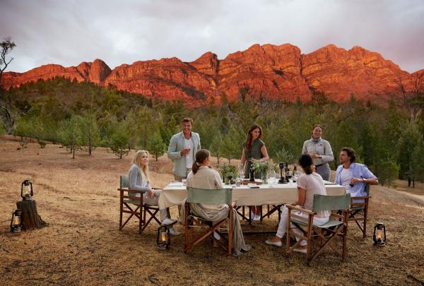 A group of guests enjoys an outdoor long-table dinner at Arkaba, Flinders Ranges, South Australia. Surrounded by lanterns on the ground, they dine as the rugged mountain range behind them glows vibrant orange at sunset. © Tourism Australia