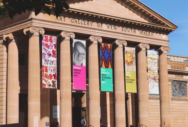 The sandstone façade of the Art Gallery of New South Wales in Sydney features grand classical columns. Vibrant exhibition banners in pink, green, and yellow hang between the pillars under a clear blue afternoon sky. © Tourism Australia
