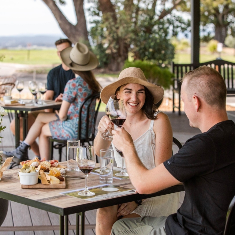 A couple enjoys a private wine tasting and cheese platter on a paved patio at Audrey Wilkinson. They are clinking glasses over a rustic wooden table, with the rolling hills and a distant dam providing a peaceful backdrop. © Agnew Wines