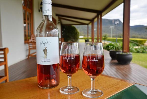 A bottle of Audrey Wilkinson rosé and two filled wine glasses sit on a wooden table on a shaded verandah. The foreground is in sharp focus, while the background reveals the sprawling estate gardens and the silhouette of the Broken Back Range in the distance. © Agnew Wines