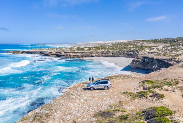 An aerial perspective of a silver 4WD vehicle parked on a rugged, rocky cliff edge overlooking the turquoise waters of the Eyre Peninsula. Two people stand near the vehicle, taking in the panoramic view of a secluded white-sand beach and the rolling waves of the Southern Ocean. © Tourism Australia 