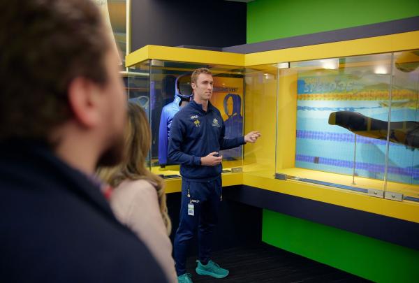 A tour guide at the Australian Institute of Sport stands in a high-tech exhibit area, gesturing toward a display case containing a sleek, black competitive rowing shell. Two visitors in the foreground listen as he explains the engineering behind Australia's elite sporting equipment. © Tourism Australia
