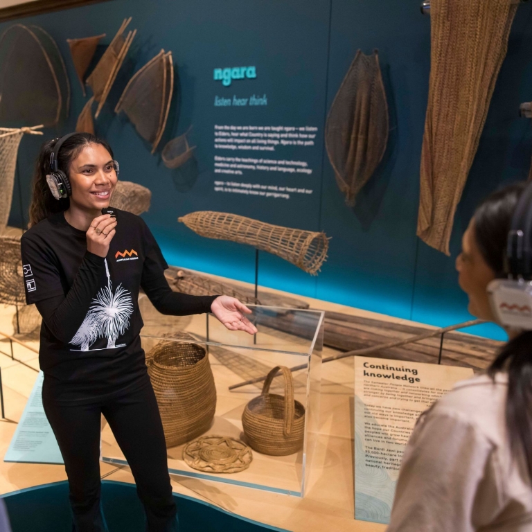 A friendly tour guide smiles warmly as they chat with a group inside The Australian Museum. The visitors listen attentively, gathered around as part of the Waranara First Nations Tour, New South Wales © Tourism Australia
