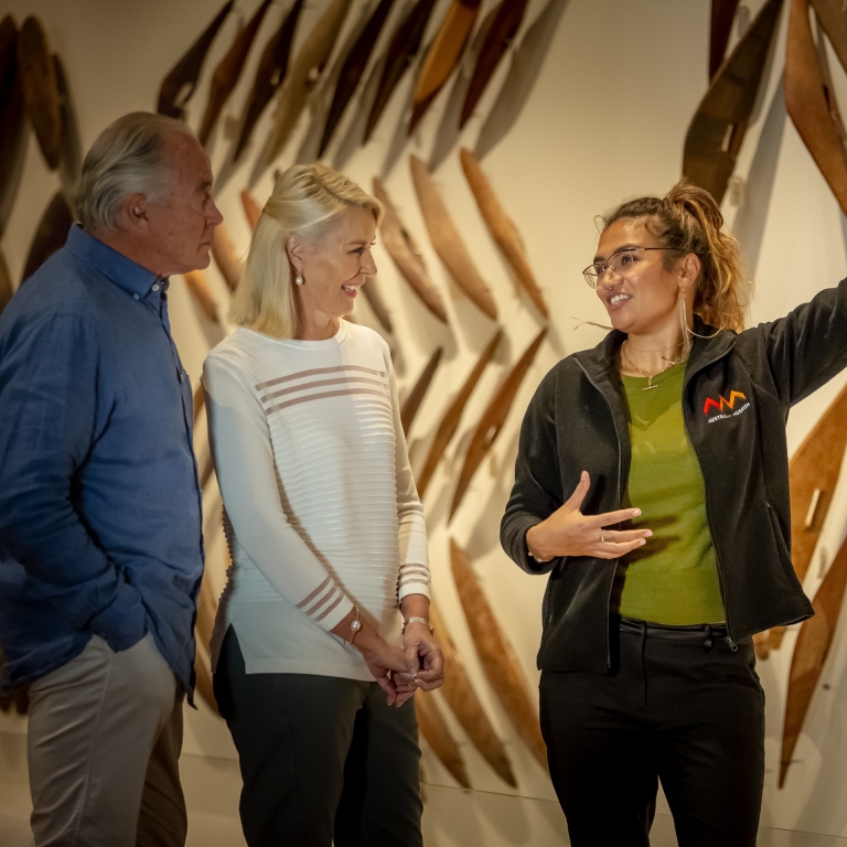 A guide presents a Waranara First Nations Tour at the Australian Museum in Sydney. She gestures toward a striking wall display of traditional boomerangs while sharing stories with an engaged couple in the New South Wales gallery. © Tourism Australia