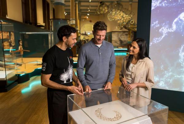 A friendly tour guide smiles warmly as they chat with a couple inside The Australian Museum. The visitors listen attentively, gathered around as part of the Waranara First Nations Tour, New South Wales © Tourism Australia