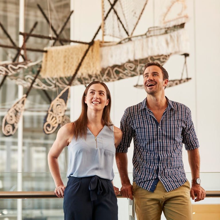 Two visitors, a man and a woman, look up with smiles at a large-scale contemporary Indigenous maritime installation. A detailed traditional outrigger boat and stylized bird figures hang suspended from the ceiling in a high-volume, sunlit gallery space. © Cultural Attractions of Australia