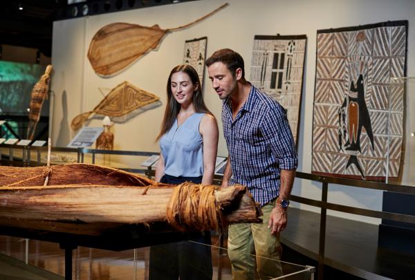 Museum visitors explore the museum's Indigenous 'Eora First People' gallery. The couple are photographed beside an Indigenous bark canoe with Saltwater bark paintings and woven art in the background. © Cultural Attractions of Australia