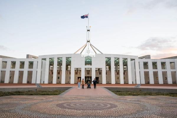 At dusk, a diverse group stands outside Australian Parliament House, with an indigenous mosaic in the foreground. The Australian flag flies overhead, and the area is disability accessible. © Tourism Australia