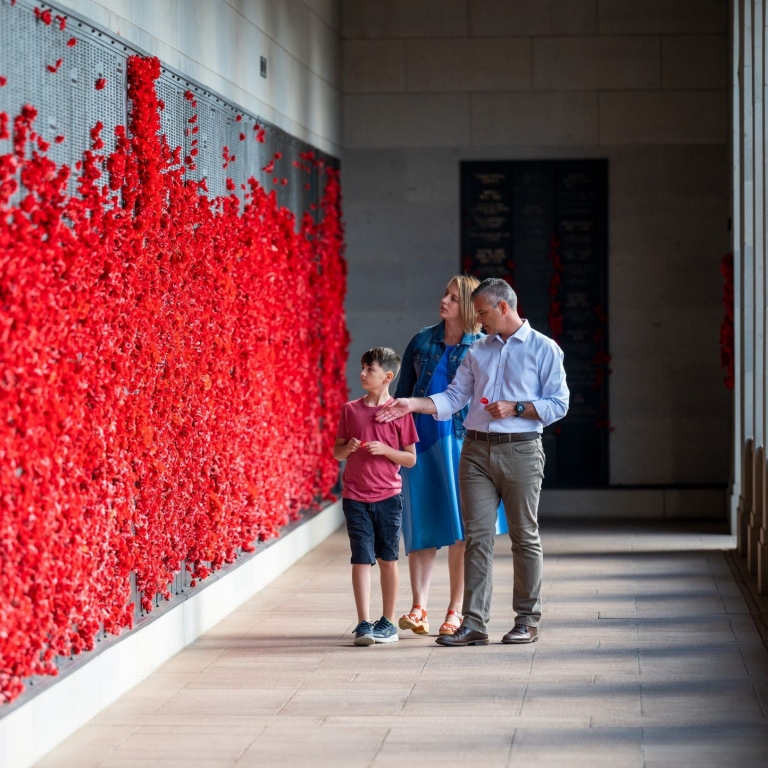 A family of three walks along the Roll of Honour, where a wall of bronze panels is covered in thousands of bright red paper poppies. A man points toward the names of the fallen, sharing a quiet moment of remembrance with a young boy and a woman. © Tourism Australia