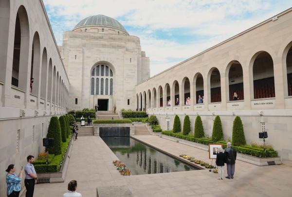 A wide view of the Australian War Memorial's Commemorative Area, featuring a long, still reflection pool leading toward the Hall of Memory with its prominent green dome. Visitors walk along the arched cloisters that house the Roll of Honour, under a bright blue sky with soft white clouds. © Tourism Australia