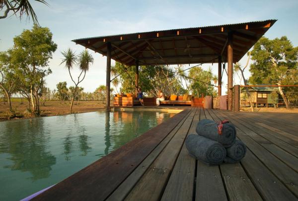 A set of three dark rolled towels sits on a wooden deck in the foreground, next to a clear, infinity-edge swimming pool at Bamurru Lodge. In the background, guests relax on comfortable lounge seating under a large open-air timber pavilion, surrounded by native trees under a soft morning sky. © Bamurru Plains