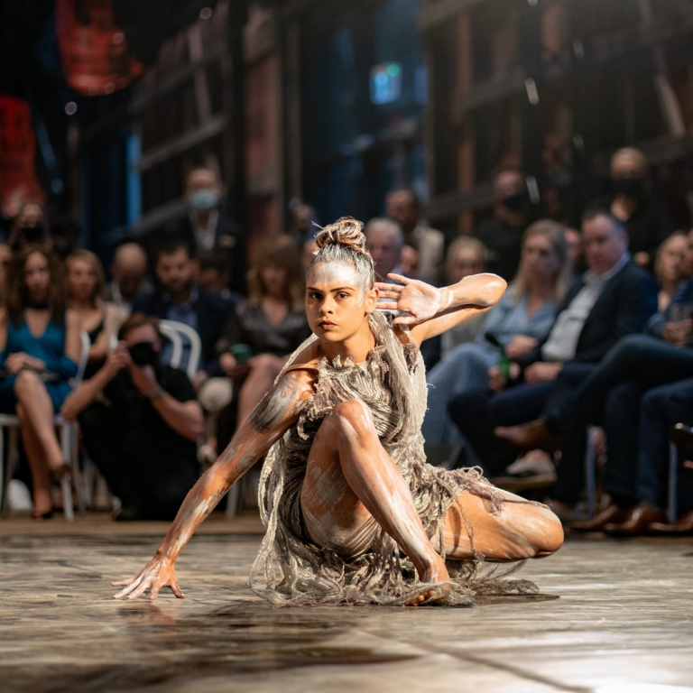 A solo female dancer, her skin marked with traditional white ochre, performs a grounded, emotive movement on a dimly lit stage. She is draped in textured, neutral-toned fabrics that flow with her posture, capturing the "fire" and spirit that the name Bangarra represents in the Wiradjuri language. © Bangarra Dance Theatre