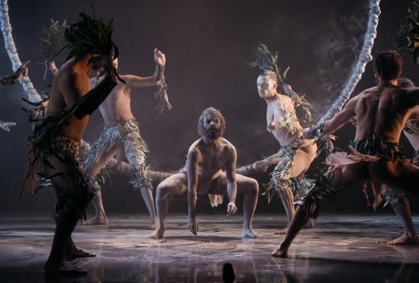 A group of male dancers performs an intense ceremonial sequence under dramatic stage lighting. In the center, a dancer crouches low with a focused expression, while others surround him with leafy branches attached to their limbs, creating a sense of ancient storytelling brought to a modern stage. © Bangarra Dance Theatre