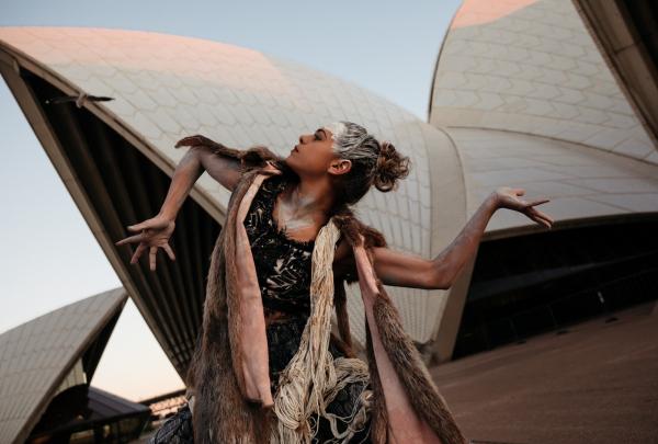 A dancer from Bangarra Dance Theatre performs a powerful contemporary piece on the forecourt of the Sydney Opera House. They wear an earth-toned costume with fur accents and traditional white body paint. Her movements dynamic and expressive against the backdrop of the iconic white sails. © Bangarra Dance Theatre