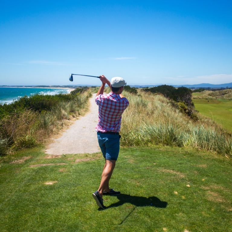 A golfer in a red checkered shirt takes a powerful swing from an elevated tee box. The rugged coastline and crashing waves of the Tasmanian coast frame the left side of the image, while the rolling dunes of the course extend to the right. © Tourism Australia