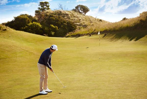 A golfer in a navy shirt and khaki trousers putts on a pristine green at Barnbougle Dunes. In the background, several wild wallabies graze peacefully on the grassy slopes of a large sand dune under a bright, clear sky. © Tourism Australia