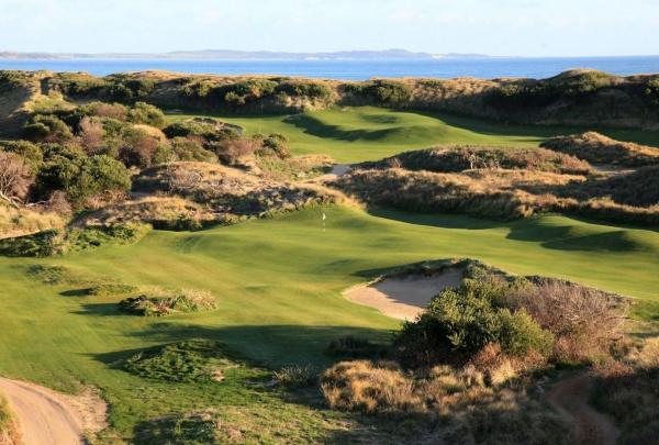 A wide landscape shot showcasing the dramatic, undulating terrain of the 18-hole championship course. Deep sand bunkers and native marram grass punctuate the vibrant green fairways, with the deep blue ocean visible in the far distance. © Links Golf Tasmania