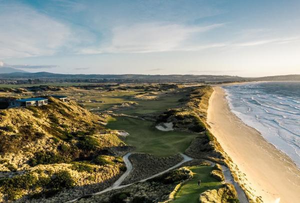 An aerial view of the rolling green fairways at Barnbougle Dunes, carved into towering coastal sand dunes. To the right, the long, white-sand beach and the turquoise waters of Bass Strait stretch toward the horizon under a soft morning light. © Tourism Australia
