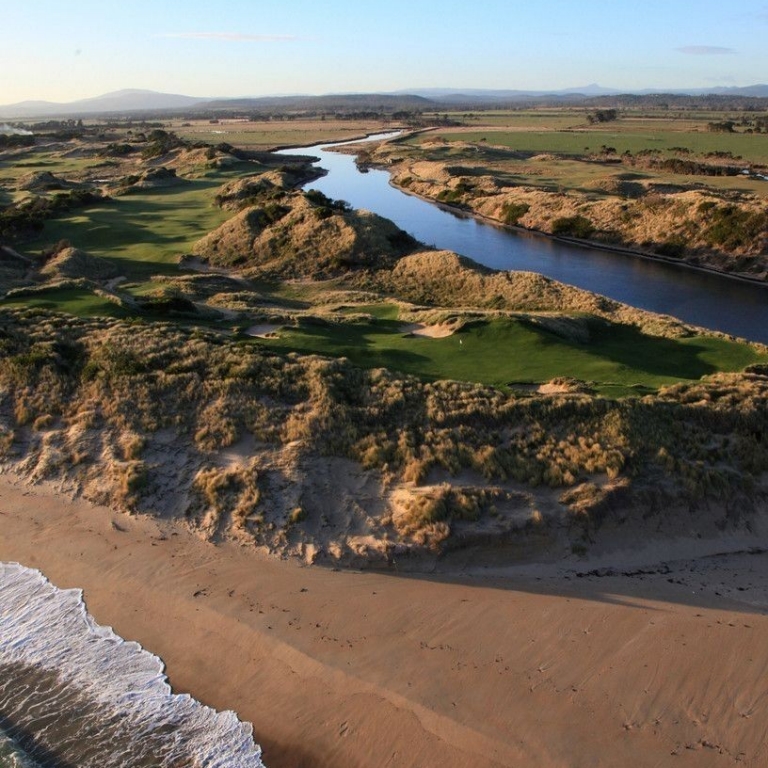 An aerial perspective of Barnbougle Lost Farm, showing a deep blue river winding through dramatic, grassy sand dunes. The vibrant green fairways of the 20-hole links course follow the natural curves of the riverbank, with the white-sand beach and rolling waves of the Bass Strait in the foreground. © Links Golf Tasmania