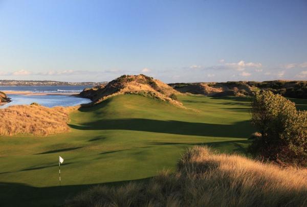 An aerial view looking inland toward the Tasmanian hinterland and distant mountains. The expansive fairway is dotted with strategic bunkers and framed by native coastal scrub, capturing the diverse routing that makes the course unique. © Links Golf Tasmania
