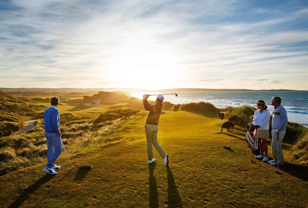 A group of four golfers stands on an elevated tee box at Barnbougle Lost Farm during a vibrant sunset. One golfer is captured in mid-swing, silhouetted against the golden light, while wild kangaroos graze peacefully on the grassy slopes nearby. © Tourism Australia