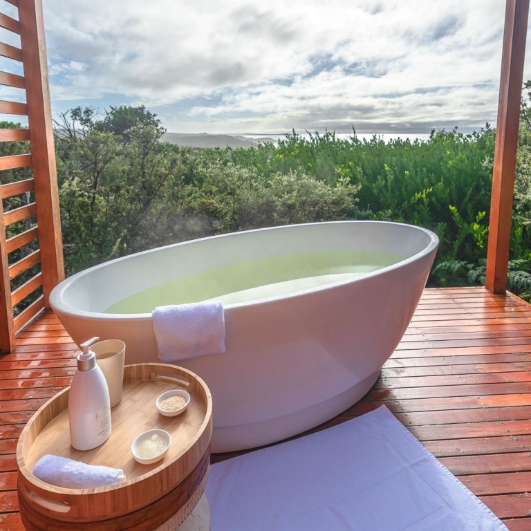 An inviting warm bath sits on a private outdoor balcony at Bay of Fires Lodge, surrounded by lush green foliage. Steam gently rises from the bathwater, suggesting perfect warmth. In the distance, the sparkling ocean is visible beyond the trees, adding to the sense of peaceful seclusion and natural luxury © Tourism Australia