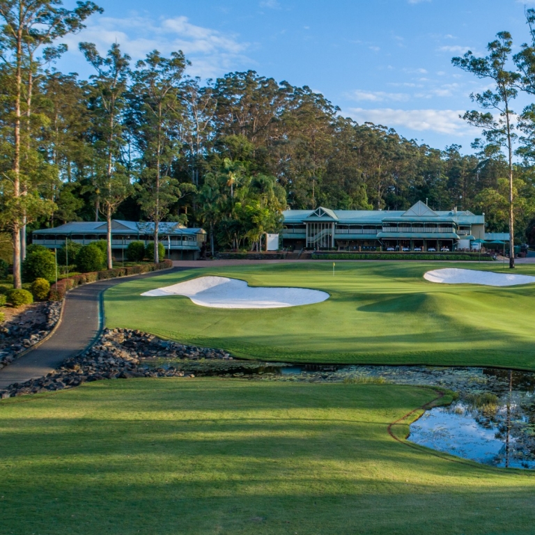 A scenic perspective of the Bonville Golf Resort clubhouse seen from across a calm water hazard. The still water in the foreground creates a mirror-like reflection of the lush green grass, the white bunkers, and the tall, slender gum trees that frame the estate. © Bonville Golf Resort