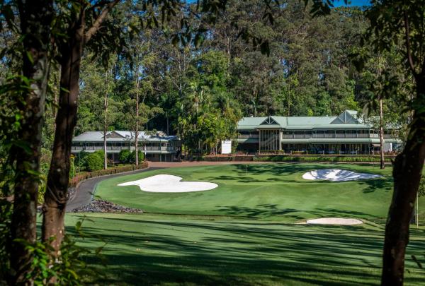 A wide view of the Bonville Golf Resort clubhouses, featuring classic green-roofed architecture nestled among a dense wall of towering flooded gums. In the foreground, a perfectly manicured green with two large, bright white sand bunkers is visible, with a paved cart path winding along the left side of the fairway. © Bonville Golf Resort