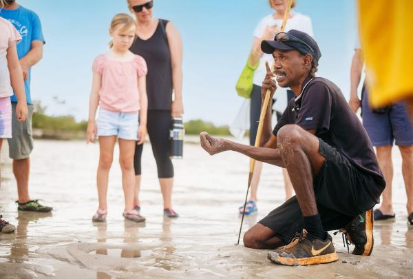 A local Bardi guide, Terry Hunter, squats on a sun-bleached tidal flat at Cygnet Bay. He holds a small object in his hand while explaining traditional coastal knowledge to a group of attentive visitors standing around him under a clear Kimberley sky. © Tourism Australia