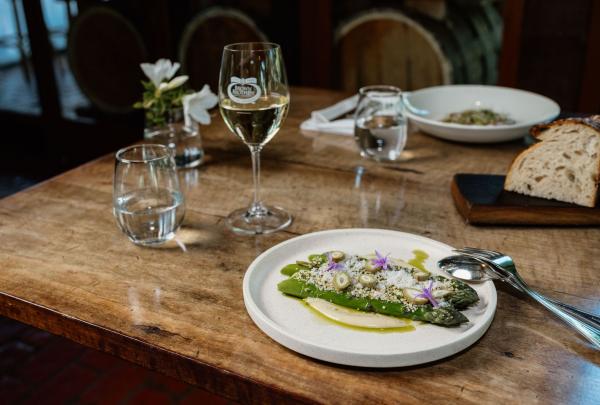 A beautifully plated dish of fresh green asparagus, topped with shaved cheese, seeds, and edible purple flowers, sits on a rustic wooden table. A glass of white wine and a plate of crusty sourdough bread accompany the meal, showcasing the winery's focus on seasonal, local produce. © Brown Brothers