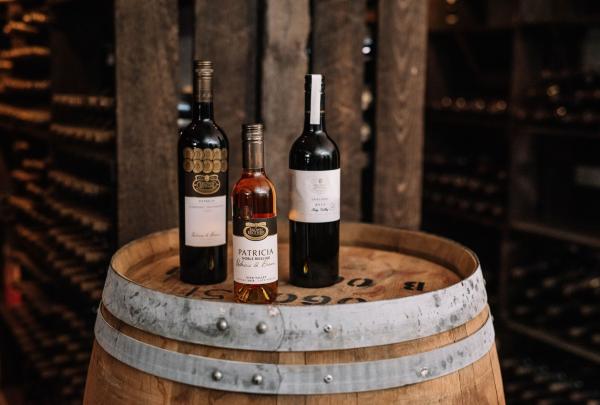 Three bottles of Brown Brothers wine—including selections from the flagship Patricia range—stand atop a weathered wooden wine barrel. The backdrop shows a blurred view of a traditional wine cellar with numerous bottles aging in dark timber racks. © Brown Brothers