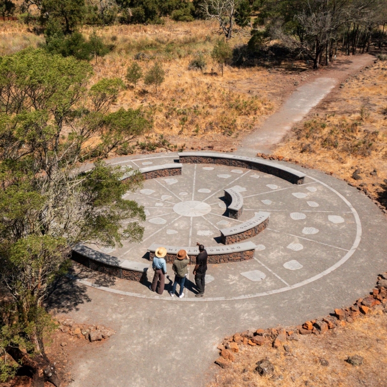 At Budj Bim Cultural Landscape Tourism on the Great Ocean Road, Victoria, an aerial view shows a circular stone gathering place. Three people stand on a sundial-like path marked with seasons, surrounded by dry grass and native trees. © Tourism Australia