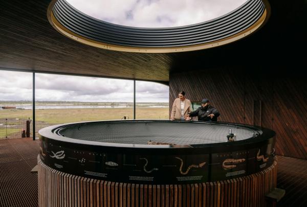 Inside the Tae Rak Aquaculture Centre, a visitor and guide look into a large circular tank. The tank's exterior features traditional illustrations and information about the lifecycle of the kooyang (short-finned eel). © Tourism Australia