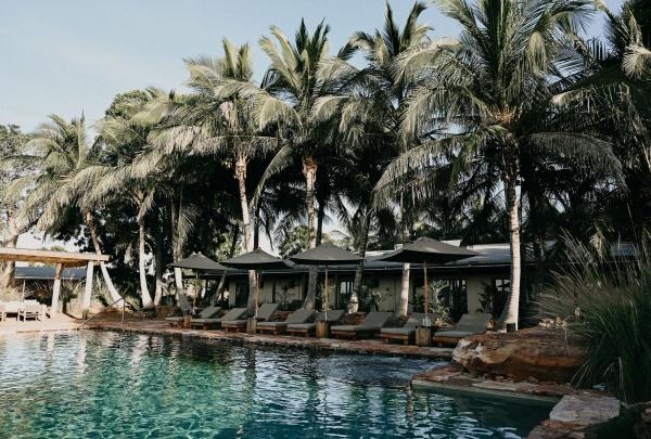 A luxury infinity pool at Bullo River Station, Northern Territory, is surrounded by tall, lush palm trees and comfortable lounge chairs with dark umbrellas. The rustic-chic station buildings are visible in the background against a clear sky. © Tourism Australia