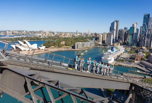 A group of climbers in grey and blue safety suits ascends the lower arch of the Sydney Harbour Bridge. From their high vantage point, they look out over a stunning panorama of Sydney Harbour, featuring the white sails of the Opera House, a large cruise ship docked at Circular Quay, and the glittering city skyline. © Tourism Australia