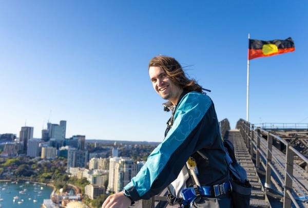 A smiling Indigenous storyteller stands on the upper arch of the Sydney Harbour Bridge during a Burrawa climb. Behind him, the Aboriginal flag flies proudly against a brilliant blue sky, with the sun-drenched suburbs of Sydney's North Shore stretching toward the horizon in the background. © Tourism Australia