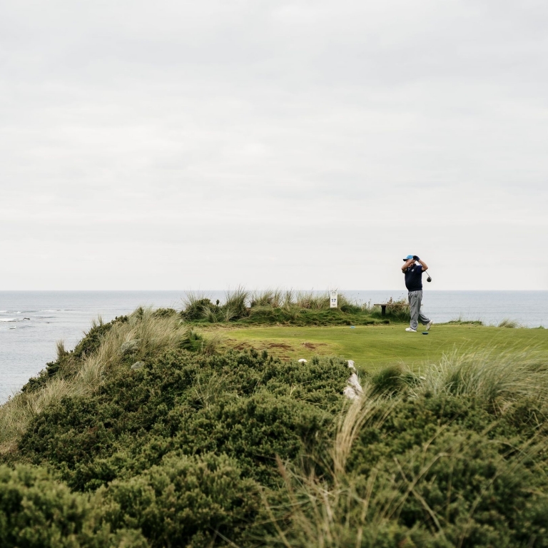 A golfer in a blue cap finishes his swing on a scenic tee box at Cape Wickham Golf Links in Wickham, Tasmania. The lush green fairway is perched on a grassy cliff overlooking the calm, grey waters of the Bass Strait. © Tourism Australia