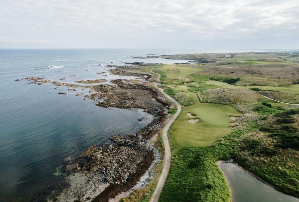 An aerial view of Cape Wickham Golf Links in Wickham, Tasmania, reveals a stunning green fairway with sand bunkers nestled alongside a rugged, rocky coastline where the deep blue ocean meets the wild Tasmanian shore. © Tourism Australia