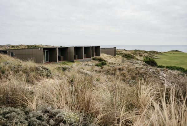 Modern, dark-grey accommodation pods at Cape Wickham Golf Links in Wickham, Tasmania, sit nestled into the grassy coastal dunes, offering guests a secluded stay with sweeping views of the green fairways and the Bass Strait. © Tourism Australia