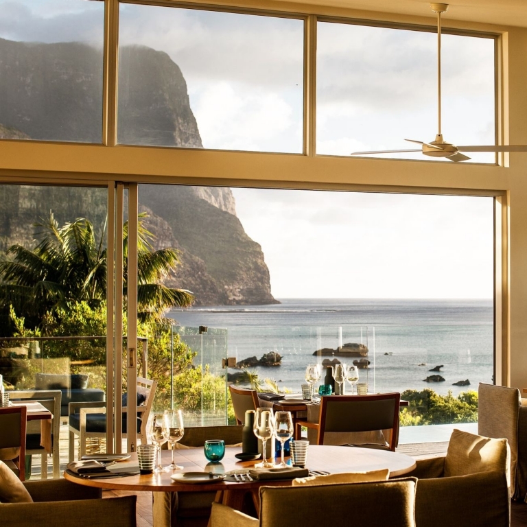 The sun-drenched dining room at Capella Lodge on Lord Howe Island features floor-to-ceiling windows that frame a spectacular view of the turquoise lagoon and the dramatic, sheer cliffs of Mount Gower under a soft afternoon sky. © Bailie Lodges