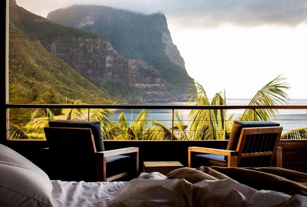 A view from a plush bed at Capella Lodge on Lord Howe Island, where two timber armchairs on a private balcony overlook swaying palm fronds and the magnificent, cloud-topped peaks of Mount Gower rising from the turquoise sea. © Bailie Lodges